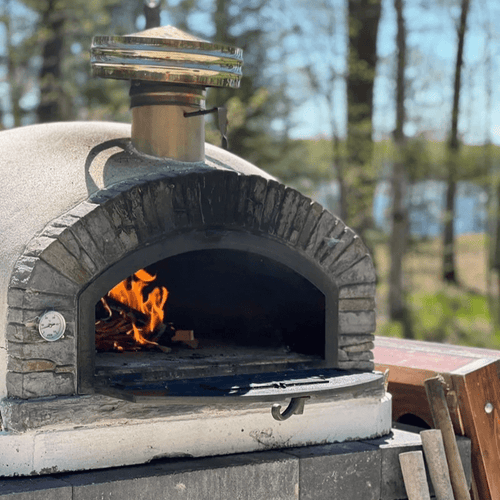 Wood-fired Buena Ventura Preto Premium Pizza Oven with flames inside, set in a forested outdoor area.